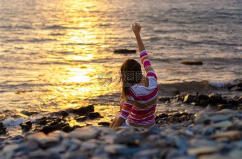 一个女孩坐在海边向水里扔石头举起手来晚上日落高清图片下载-正版图片307056154-摄图网