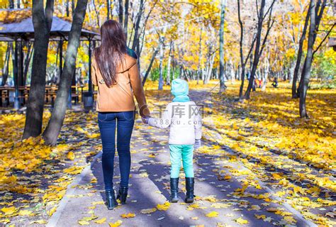 在温暖的阳光明媚的日子里，年轻母亲和她可爱的女儿在黄色的秋天森林里散步的背影高清摄影大图-千库网