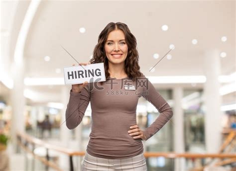 销售、就业和商业概念-快乐的女售货员或女售货员在购物中心背景上挂着招聘横幅。高清摄影大图-千库网