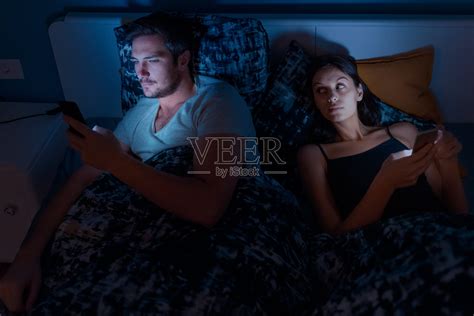 夫妻晚上在床上玩手机，女人用怀疑的目光看着照片摄影图片_ID:351217176-Veer图库