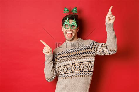 戴着派对眼镜和圣诞毛衣的快乐帅哥的画像，一边跳舞一边指指点点，享受新年庆祝活动，站在红色背景上高清摄影大图-千库网