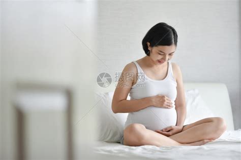 愉快腹部怀着白帕贾马斯的美丽亚洲孕妇身穿白色睡袍坐在床上微笑着健康快乐的年轻母亲在怀孕时抱着摸和看肚子在家放松高清图片下载-正版图片 ...