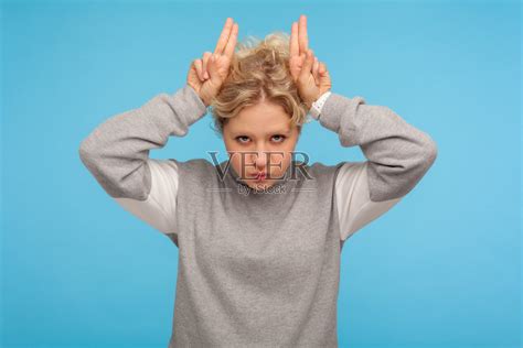 咄咄逼人的女人在灰色运动衫显示牛角手势与手指在头上和鼓胀的脸颊照片摄影图片_ID:349862000-Veer图库