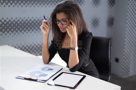 身着黑色西装戴着昂贵手表和眼镜的漂亮黑发美国黑人女商人坐在办公室的桌子旁高清图片下载-正版图片503359537-摄图网