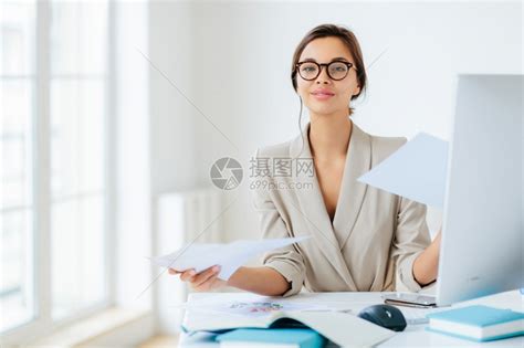 工作繁忙的女经理在办公室工作有文件在办公室工作桌面上摆姿势佩戴眼镜和正规服装忙于编写报告自信地看相机高清图片下载-正版图片303688805-摄图网