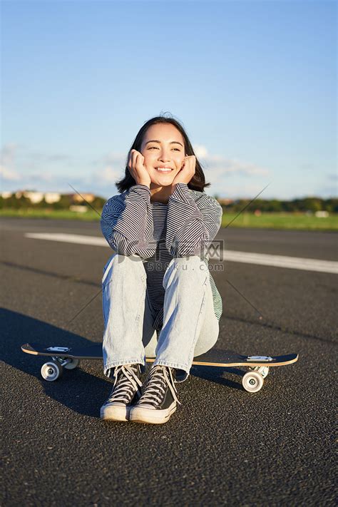 亚洲女滑手的垂直镜头，坐在她的滑板上微笑，享受阳光明媚的日子，在户外空荡荡的路上在长板上巡航高清摄影大图-千库网